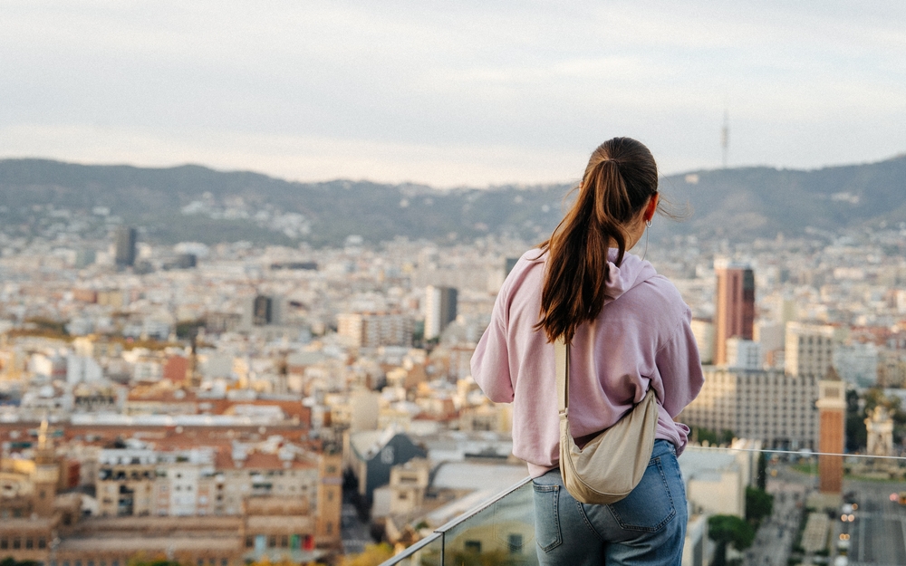 A woman with a pony tail overlooks the cityscape in her pink hoodie, jeans, and crossbody bag.