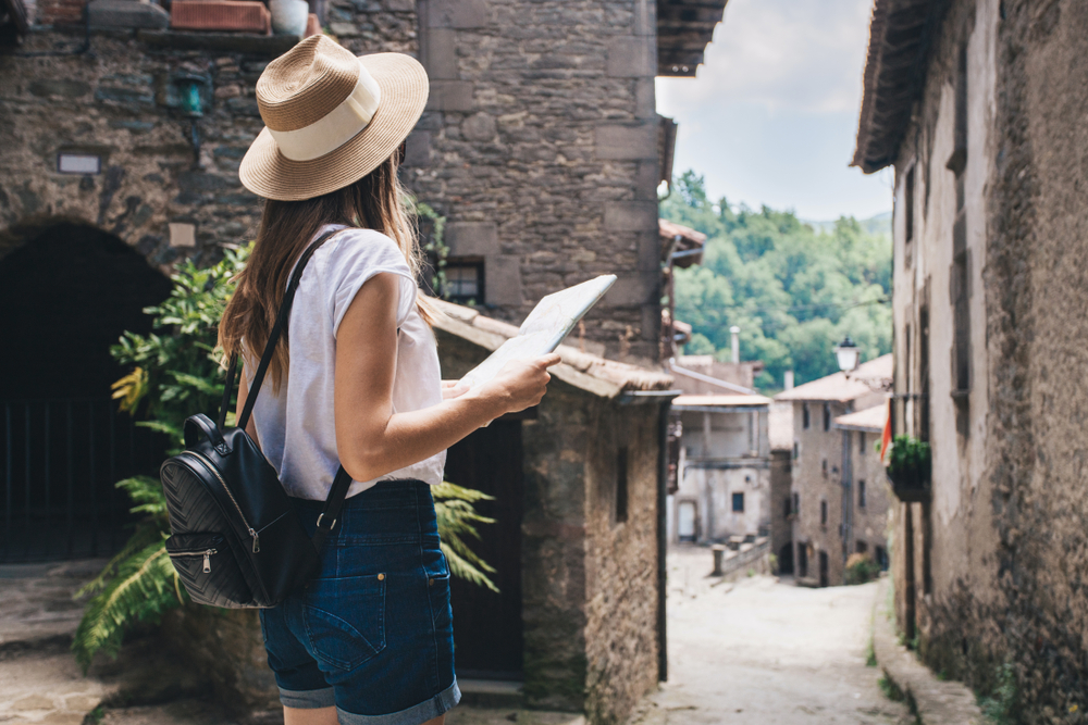A woman pauses to look at a map and then her surroundings, wearing a white top, jean shorts, a backpack and a straw hat.