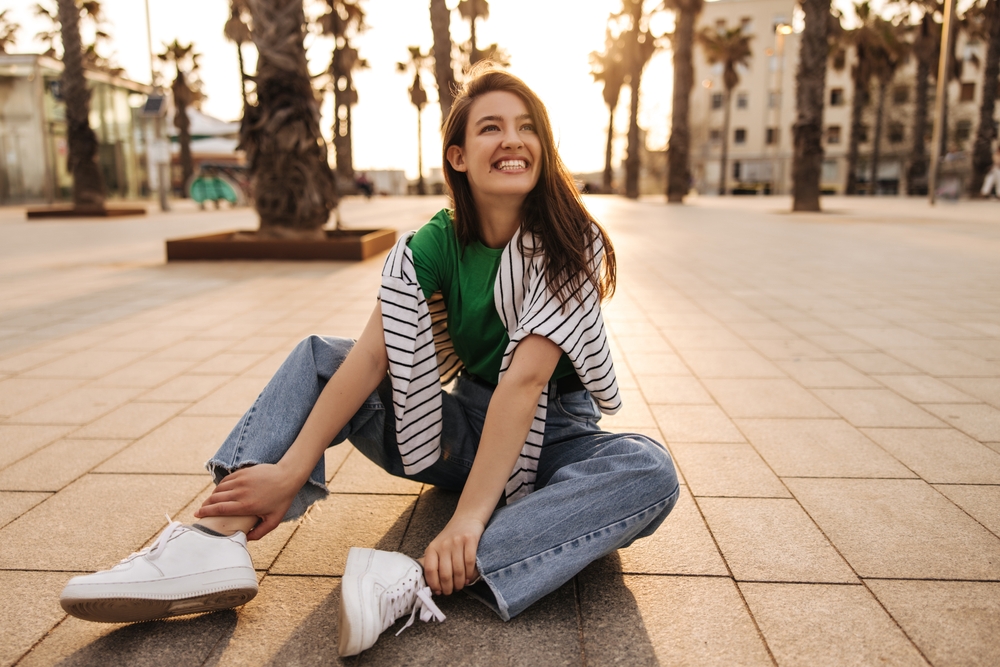 A woman sits on the ground of a city in jeans, white sneakers, a green top, and a sweater over her shoulders. She smiles at the camera.