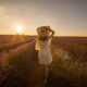 A woman walks down a row of purple flowers at sunset, wearing a white sundress and sunhat, perfect for what to wear in south France.