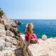A curly hair blonde sits on rocks overlooking the ocean, while wearing a pink maxi dress, confident in what she knew about what to wear in the French Riviera.