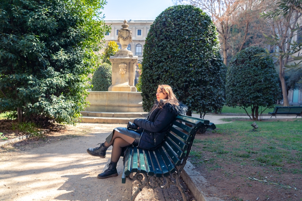 A woman knows what to wear in Spain as it gets colder: she sits on a bench by a fountain, clad in a leather skirt, boots, tights and a parka.