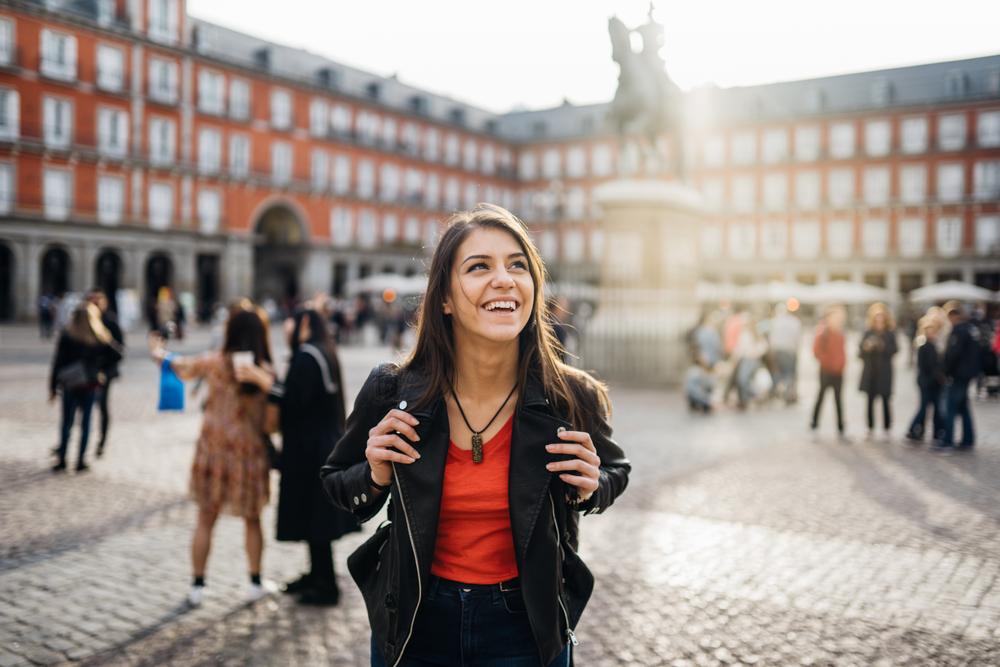 A young brunette looks up smiling at a town square while waring jeans, a red v-neck, and a leather jacket. Her hands sit at the straps of her backpack.