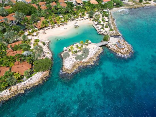 drone view of Baoase luxury resort Curacao which features rocky coastline and buildings with red and orange roofs surrounding a beach in a cove