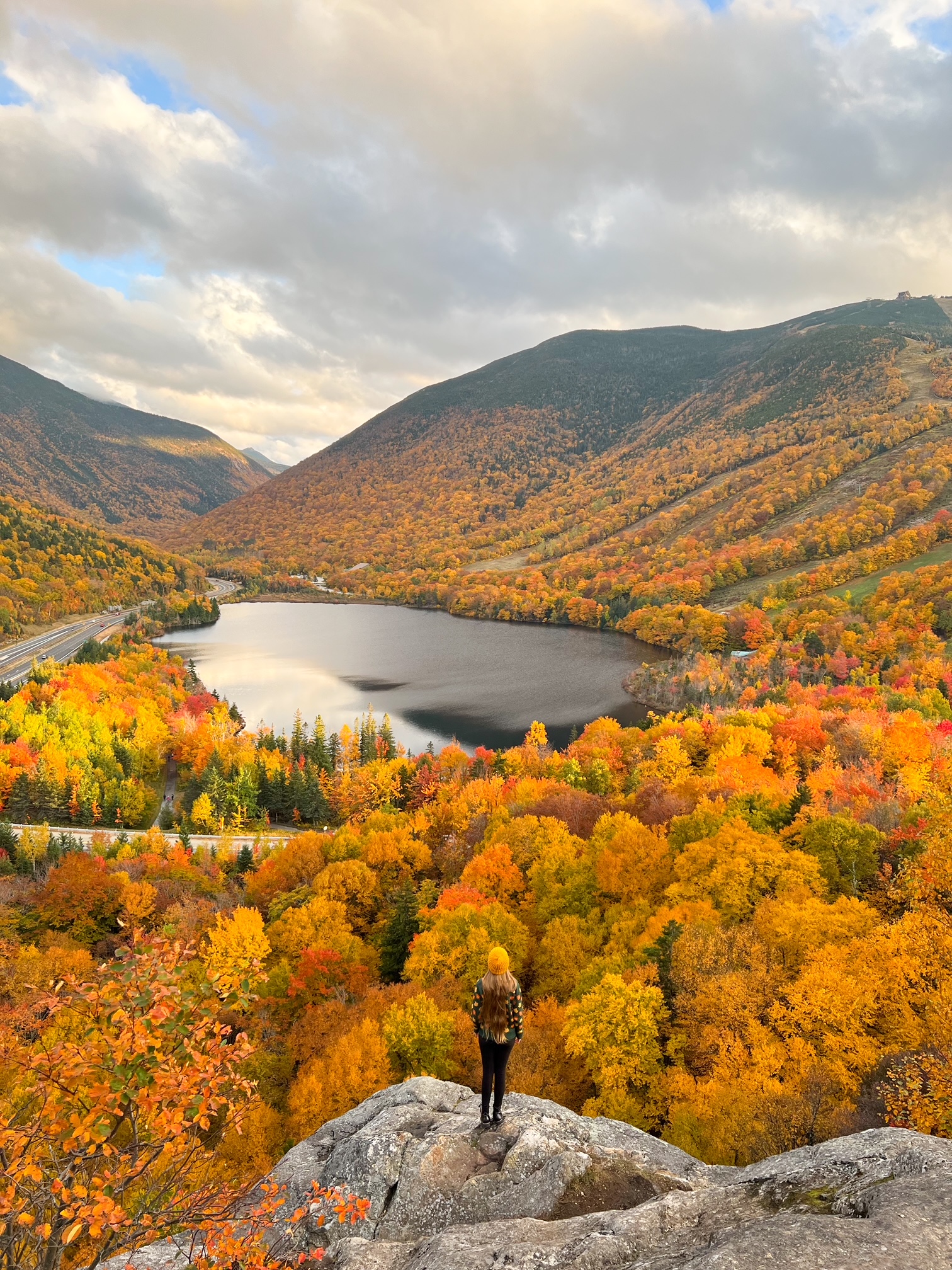 A woman in an orange beanie and sweater stands on the cliffside over yellowing and orange trees!