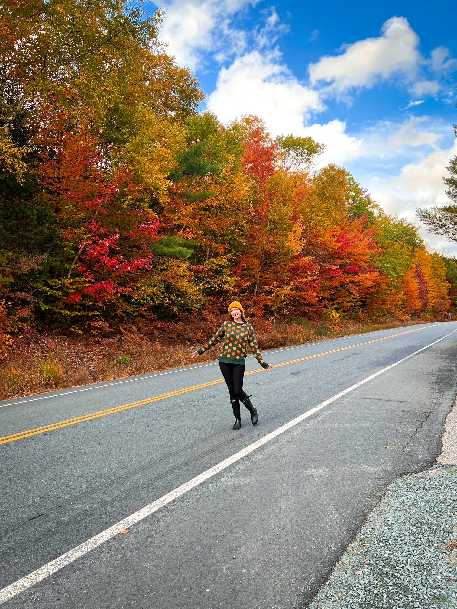 A woman in an orange beanie and leaf sweater smiles in the middle of the road as the trees turn around her, and she knows what mistakes to avoid on a fall foliage trip.