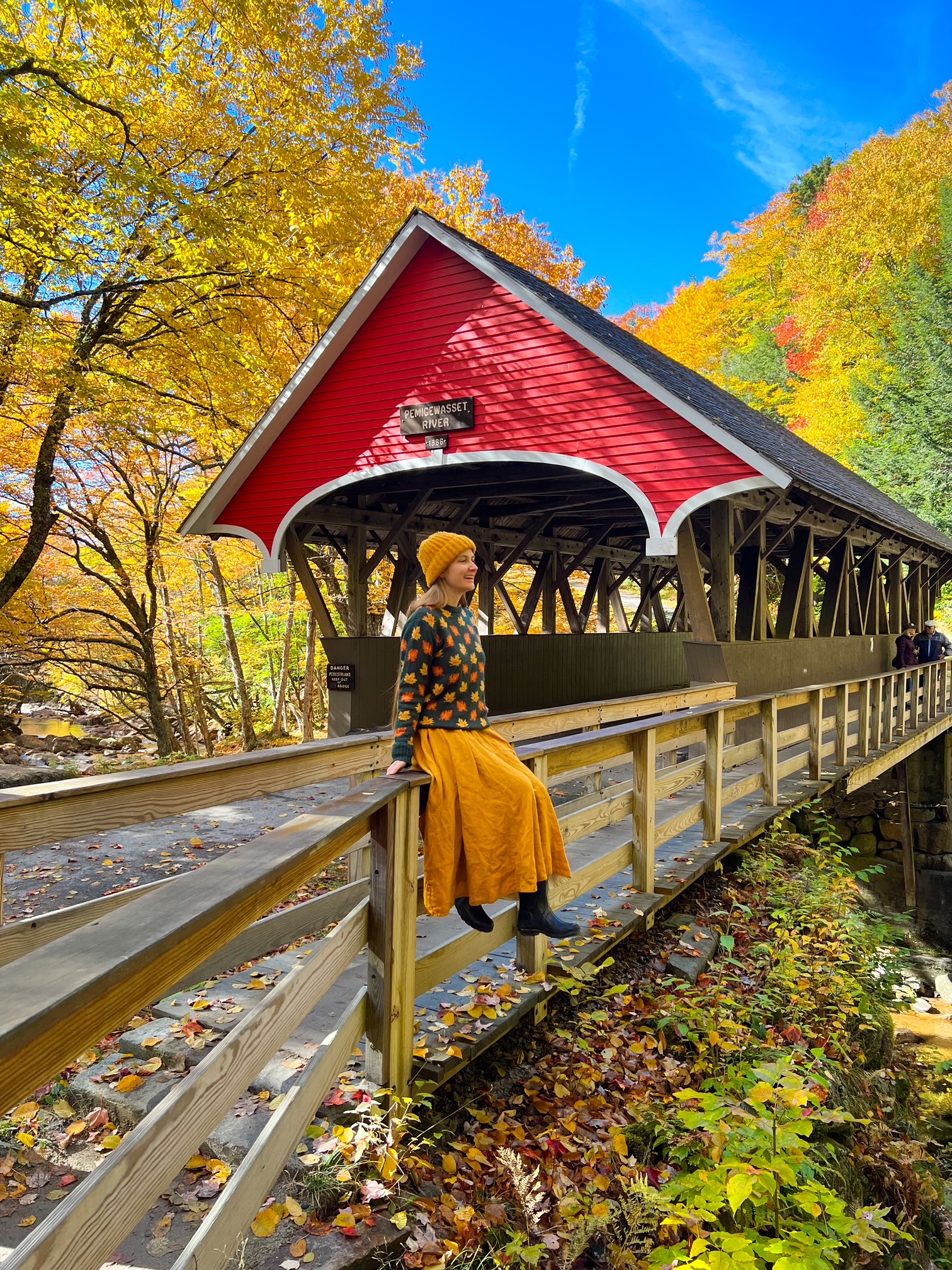 A red covered bridge crosses the Pemigwasset river, and a girl sits on the edge with the trees turning around her: she laughs as she knows the mistakes to avoid on a fall foliage trip.
