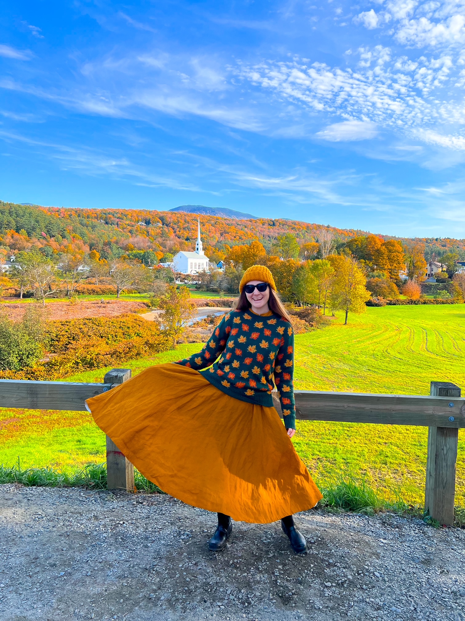 A woman in an orange skirt, beanie, and leaf sweater smiles on a road with a white church in the background, and trees turning orange beyond her.