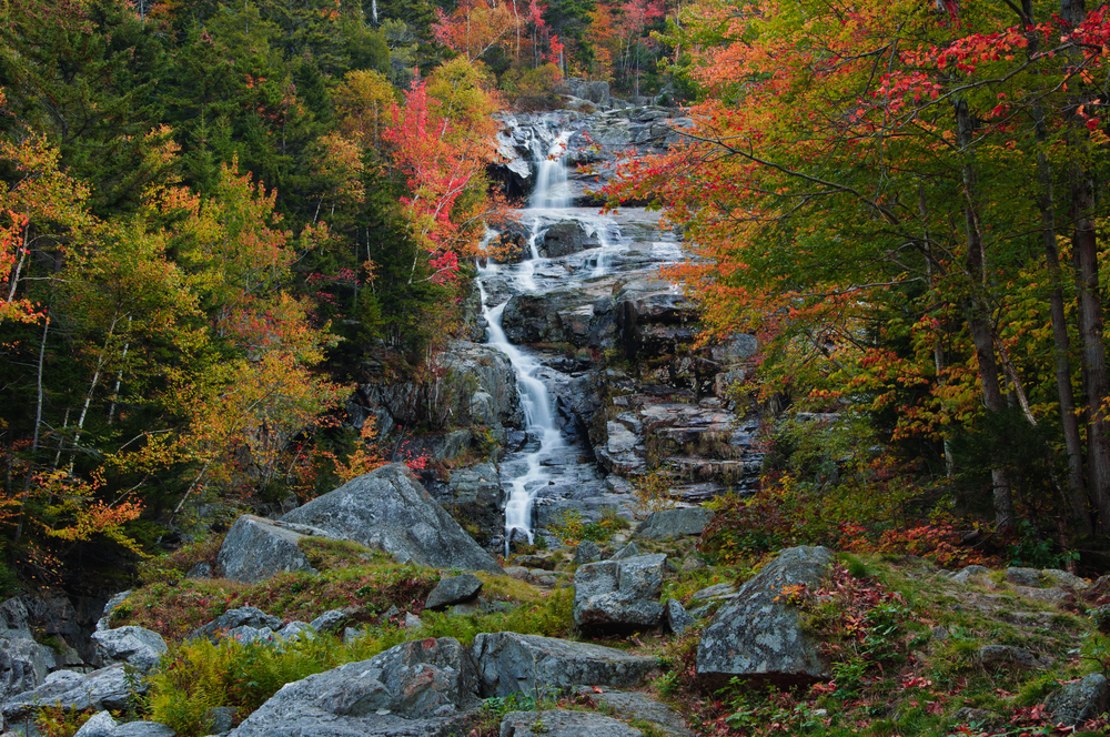 A waterfall cascades down dark rocks as the trees around it turn different shades of orange and red: skipping things like this is one of the main mistakes to avoid on a fall foliage trip.