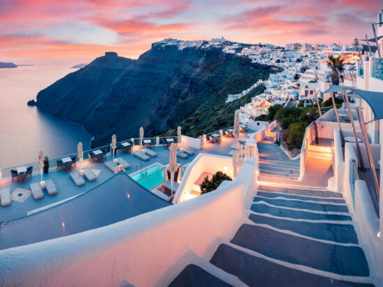 grey staircase in greece with mountains in the background and white buildings all around on the right. to the left there is water and it is sunset. the sky is pink with some pink clouds