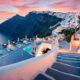 grey staircase in greece with mountains in the background and white buildings all around on the right. to the left there is water and it is sunset. the sky is pink with some pink clouds