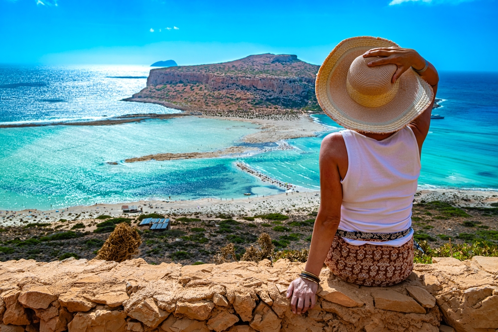 A woman overlooks the blue waters and sandy beaches in a tank and straw hat in Greece.