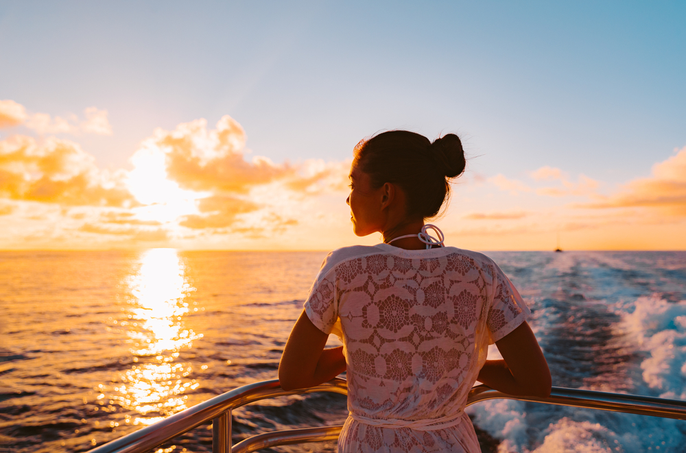 A woman overlooks the water on a ferry while the sun sets.