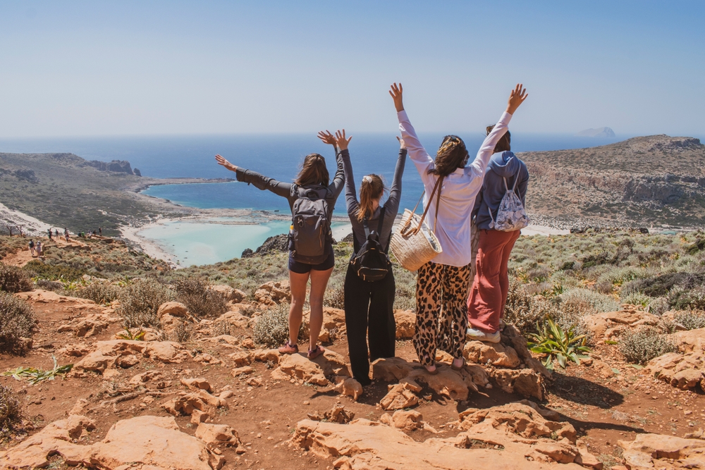 A group of friends overlook blue waters and beaches while on the cusp of a hike, their hands in the air, enjoying their trip because they knew mistakes to avoid in Greece.