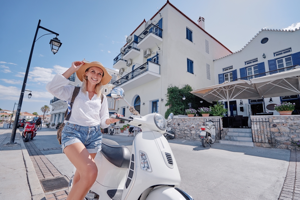 A woman in a town sits by a scooter while wearing a straw hat, a white shirt and jean shorts: she knows how to drive the scooter, which is one of the mistakes to avoid in Greece.
