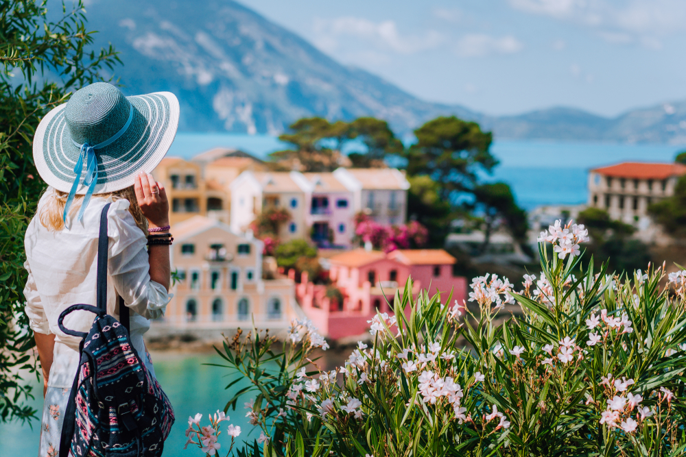 A woman in a large blue hat and white shit and backpack overlooks the small, colorful town below her, knowing which mistakes to avoid in Greece.