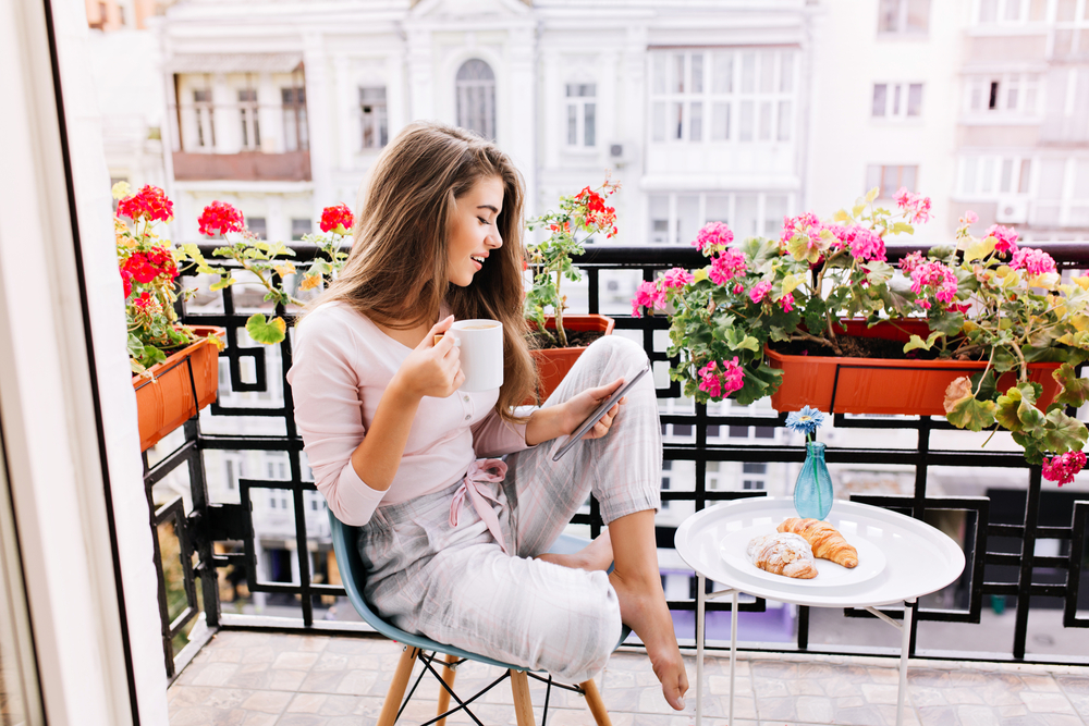 A young girl sits on the edge of her apartment balcony and scrolls her phone while drinking coffee, relaxing, since she did not overbook herself and knew what mistakes to avoid in Paris.