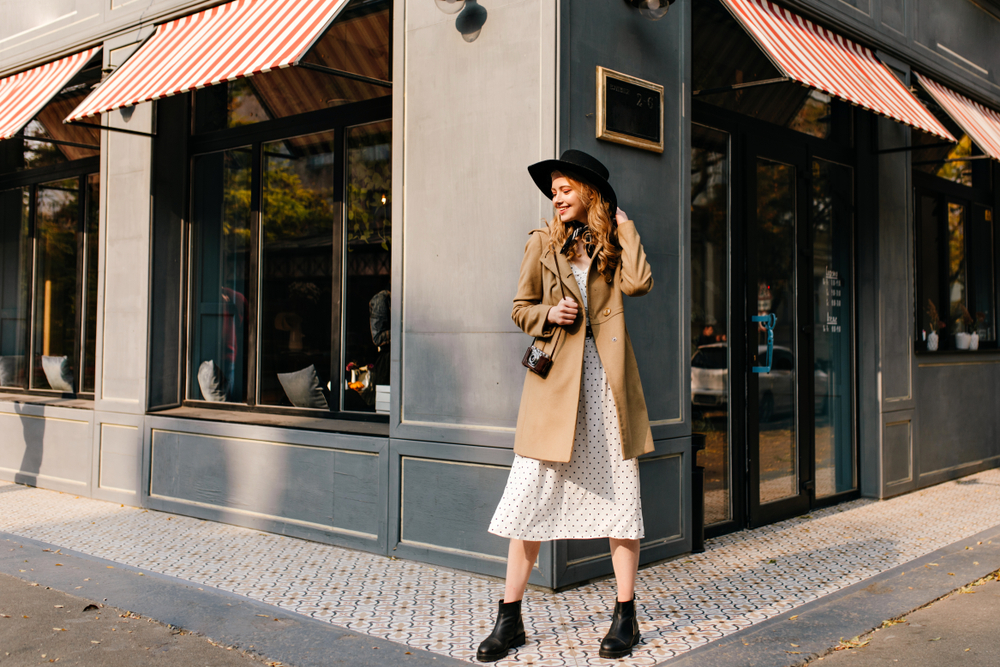 A woman in a polka dot dress, boots and trench coat smiles at the corner of a Paris shop, enjoying the city.