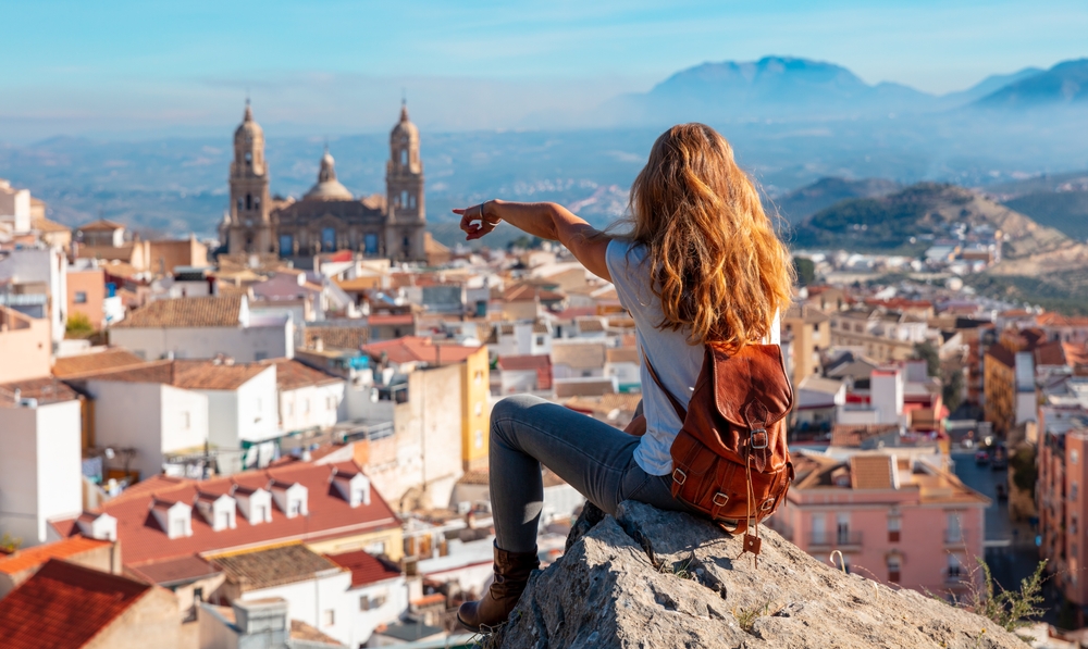 A young blonde sits on the edge of a rock and points at the town below her.