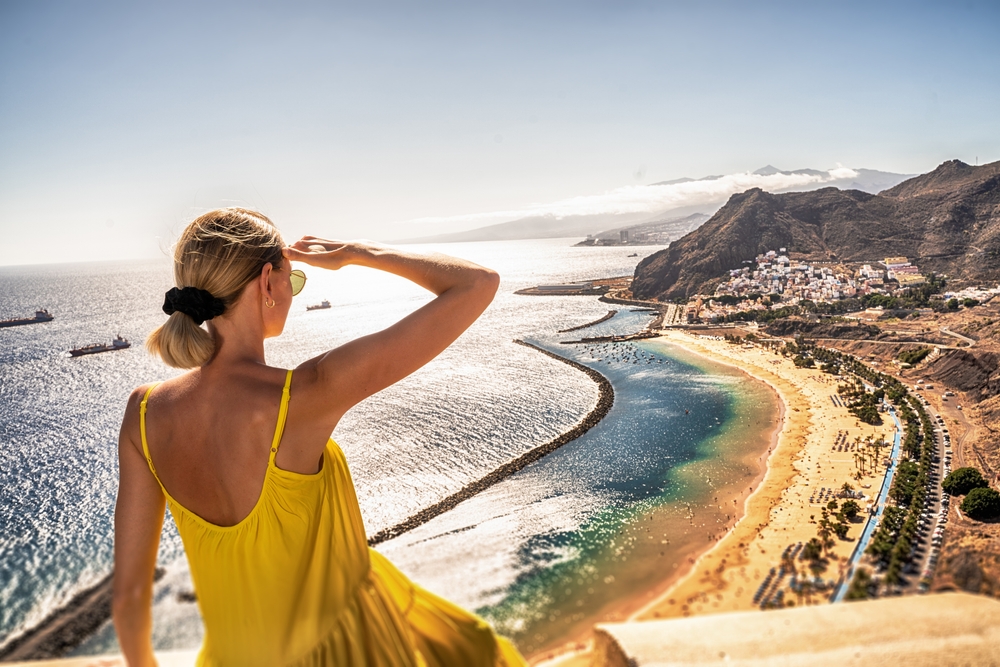 A woman in a yellow sundress overlooks the beaches in Spain, her sunglasses and hair pulled back as she looks.
