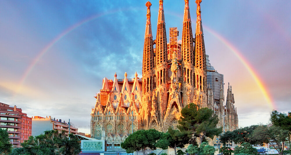 giant church in a city with a rainbow behind it. the church has multiple spires and is brown. there are green trees in the foreground and there is a pink and blue slightly cloudy sky with a rainbow going behind the church