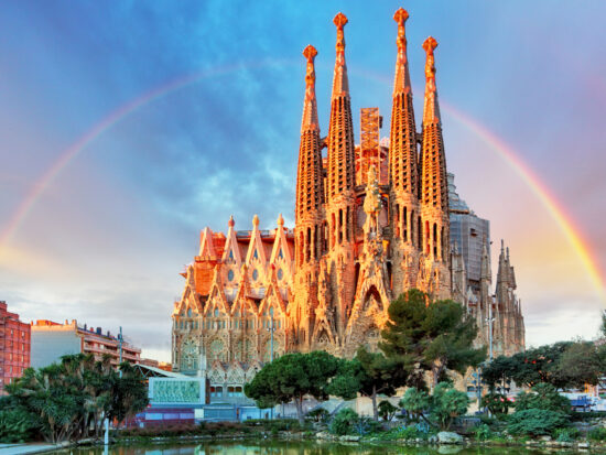 giant church in a city with a rainbow behind it. the church has multiple spires and is brown. there are green trees in the foreground and there is a pink and blue slightly cloudy sky with a rainbow going behind the church