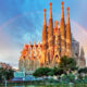 giant church in a city with a rainbow behind it. the church has multiple spires and is brown. there are green trees in the foreground and there is a pink and blue slightly cloudy sky with a rainbow going behind the church