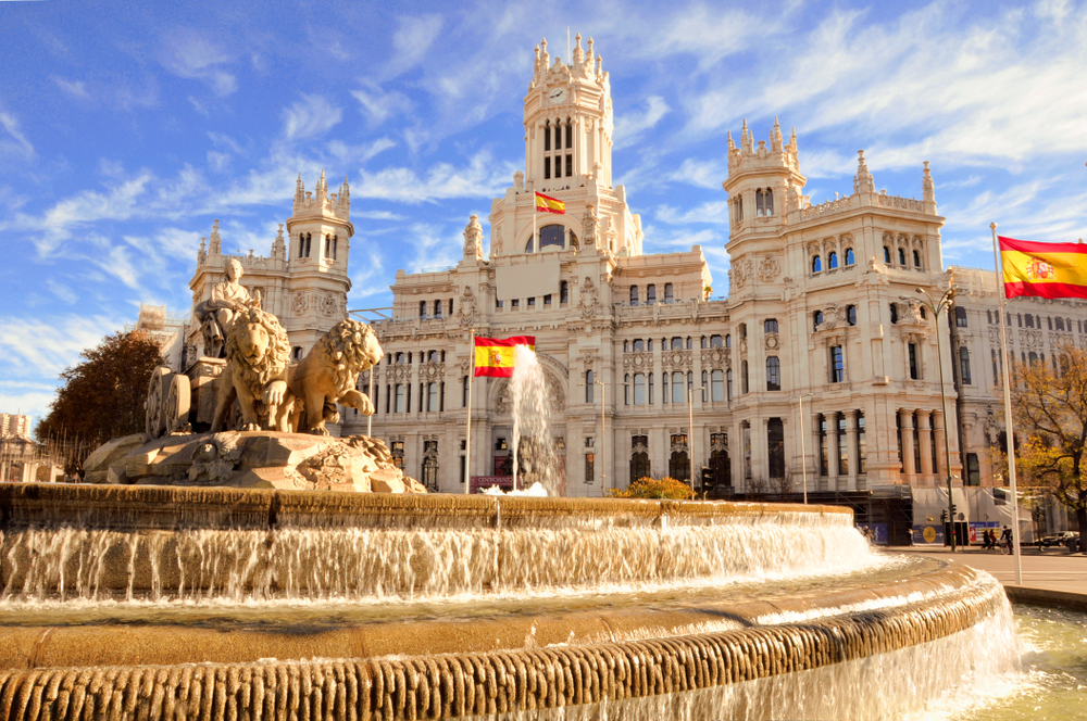 A castle and fountain stand bright in the sunlight with Spain's flags billowing overhead, encouraging you to visit Spain but to know what mistakes to avoid in Spain.