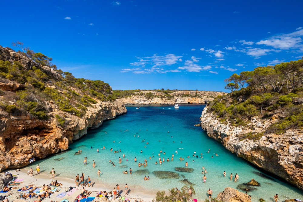 An ariel shot shows a blue lagoon type area in Spain as guests swim in the blue waters and enjoy tanning on the beach in between rocky terrain.