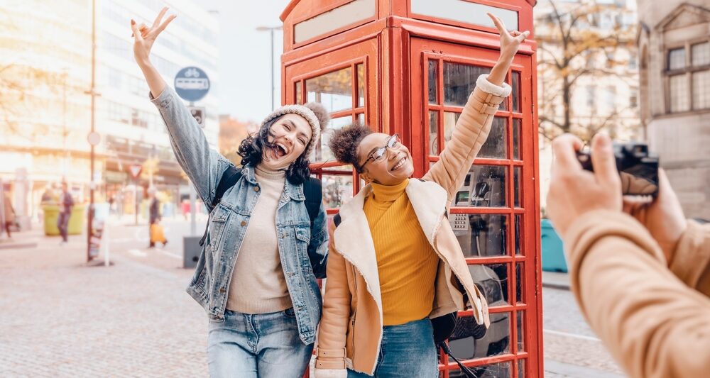 Two girls taking a picture in front of red telephone boxes. one girl is wearing jean jacket and jeans one girl is wearing yellow shirt with jacket and jeans. someones hands are in the picture taking a photo of the two girls. The article is on planning a trip to London.