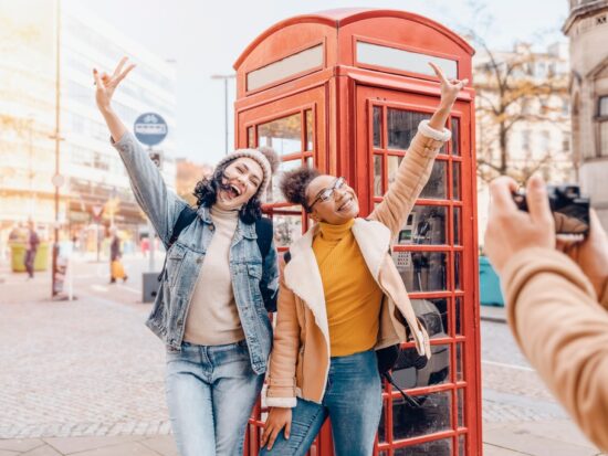 Two girls taking a picture in front of red telephone boxes. one girl is wearing jean jacket and jeans one girl is wearing yellow shirt with jacket and jeans. someones hands are in the picture taking a photo of the two girls. The article is on planning a trip to London.