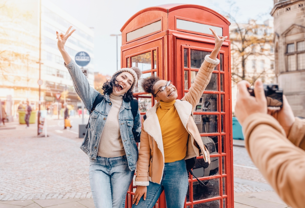 Two girls taking a picture in front of red telephone boxes. one girl is wearing jean jacket and jeans one girl is wearing yellow shirt with jacket and jeans. someones hands are in the picture taking a photo of the two girls. The article is on planning a trip to London.