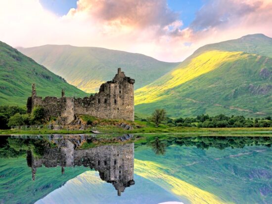 Beautiful Kilchurn Castle on Loch Awe in the highlands of Scotland at sunset with reflections. The castle itself is brown and the water is calm like glass with a reflection and the background is green hills and a blue sky with pink clouds. The article is about Scotland mistakes to avoid.