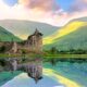Beautiful Kilchurn Castle on Loch Awe in the highlands of Scotland at sunset with reflections. The castle itself is brown and the water is calm like glass with a reflection and the background is green hills and a blue sky with pink clouds. The article is about Scotland mistakes to avoid.