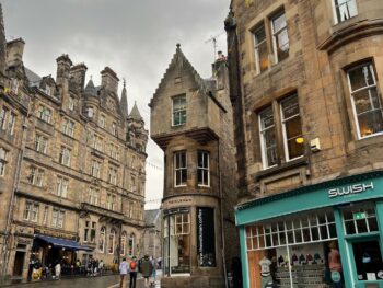 streets of Edinburgh with cobblestone street and brown brick stores. there are people walking outside of the stores. one storefront is green with brown on top with the words "swish" on top of it