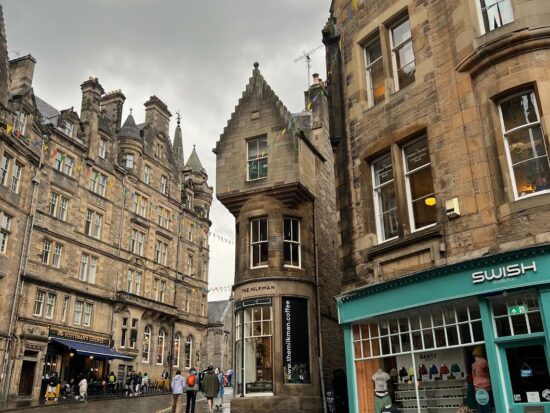 streets of Edinburgh with cobblestone street and brown brick stores. there are people walking outside of the stores. one storefront is green with brown on top with the words "swish" on top of it