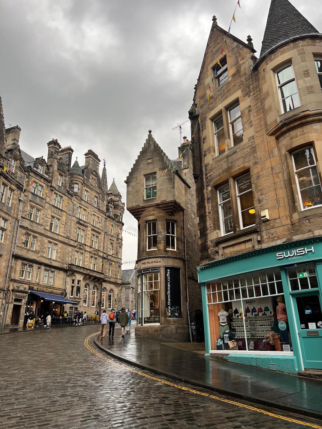 streets of Edinburgh with cobblestone street and brown brick stores. there are people walking outside of the stores. one storefront is green with brown on top with the words "swish" on top of it