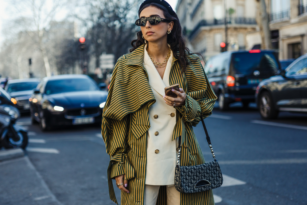 A woman makes a bold statement as she walks down the street, wearing a white trench coat with a bright colored pattern poncho over it.