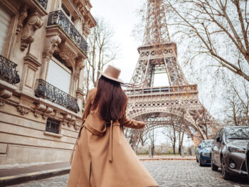 A woman in a wool trench coat looks at the Eiffel tower from a cobblestone street, while wearing a matching suede tan hat. Even her ankle boots match: this is how you know what to wear to Paris in November.