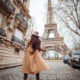 A woman in a wool trench coat looks at the Eiffel tower from a cobblestone street, while wearing a matching suede tan hat. Even her ankle boots match: this is how you know what to wear to Paris in November.