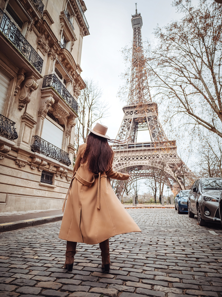 A woman in a wool trench coat looks at the Eiffel tower from a cobblestone street, while wearing a matching suede tan hat.