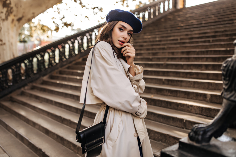 On a set of stairs a woman looks over her shoulder in her tan trench coat, blue beret, and crossbody bag.