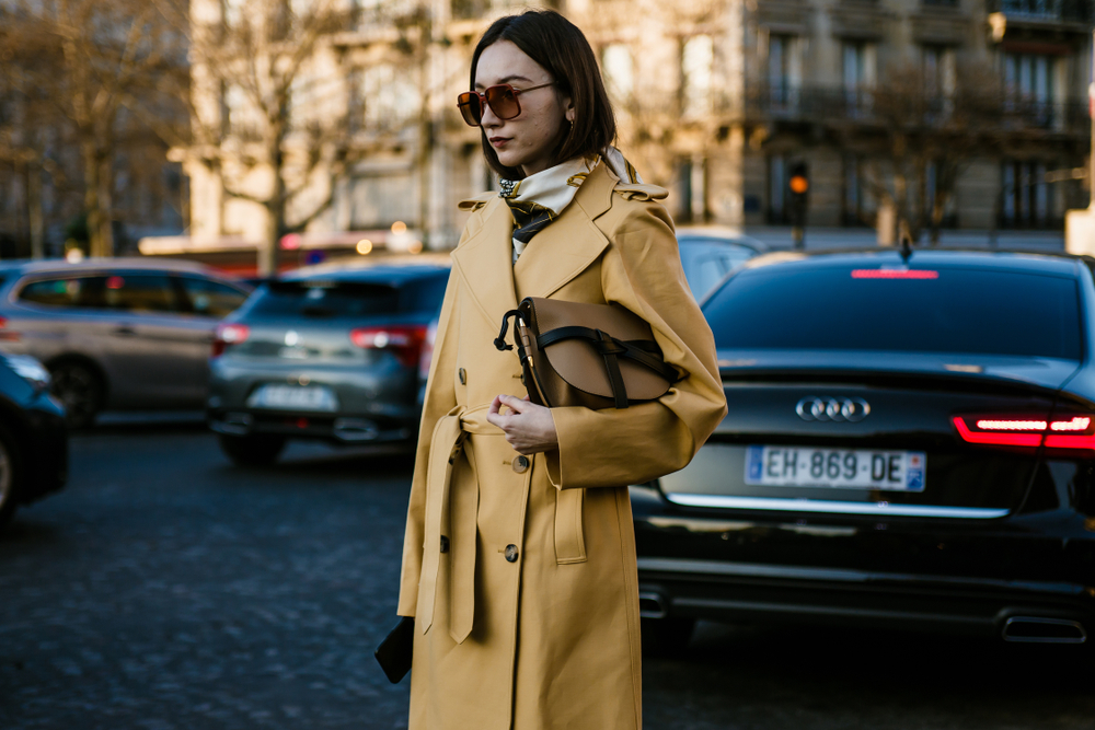 A woman crosses the street in Paris with a crossbody bag, a yellow trench coat, and a silk scarf.
