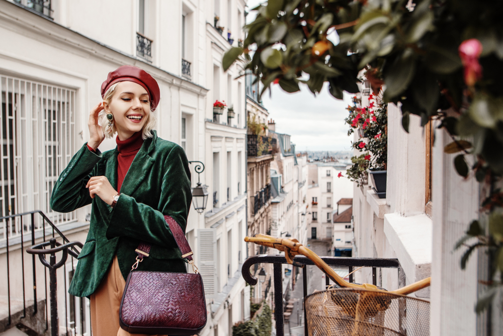 A woman knows What to wear to Paris in November as she stands on stairs above the city in a green blazer, red turtleneck, and statement fedora!