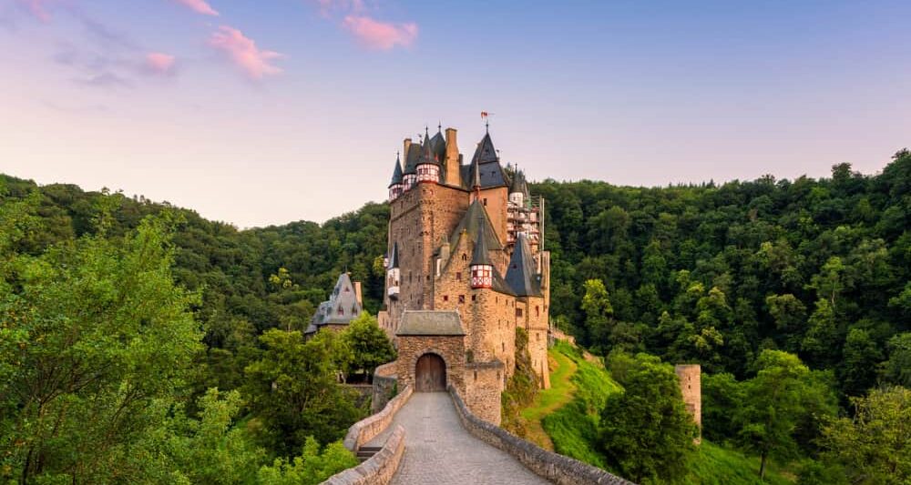 the Eltz castle that at sunset with the castle surrounded but the forest is Eltz. the castle is brown with grey roof and a pink cloudy sky