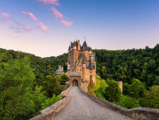 the Eltz castle that at sunset with the castle surrounded but the forest is Eltz. the castle is brown with grey roof and a pink cloudy sky