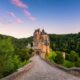 the Eltz castle that at sunset with the castle surrounded but the forest is Eltz. the castle is brown with grey roof and a pink cloudy sky