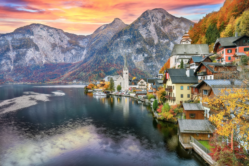 Mountains tower in the background of this stunning photo: you can go visit places like this when you prep with the things to buy from Amazon for Europe: the coastal town is bright with the turn of leaves, and they match the autumn hues of the homes and church on the water, with the sun in the back.