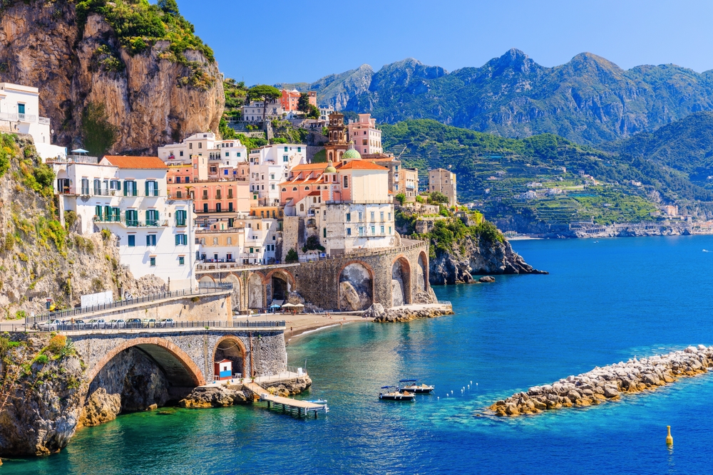 On the Italian coast, tall white and orange buildings lean against the mountainside, and tall cobblestone bridges arch over blue water.