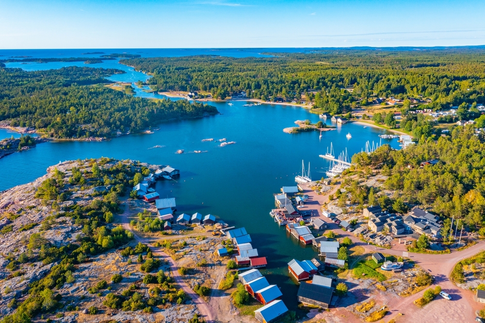 An overhead shot shows green trees surrounding blue waters in coastal inlet, with boats and docks and little homes staggered in between.